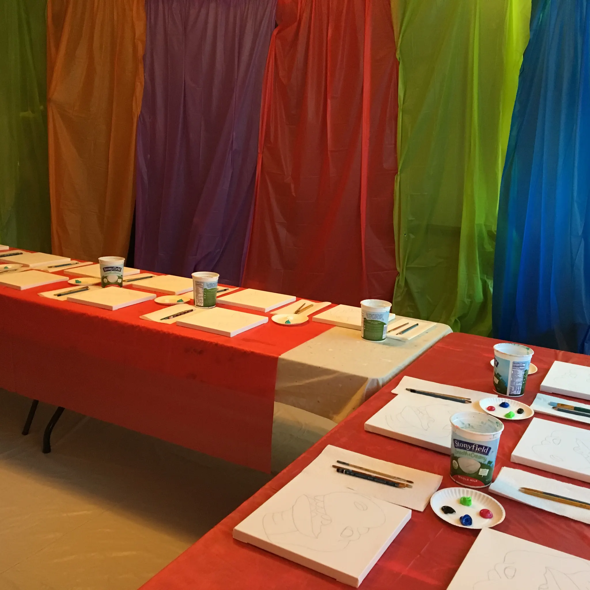 Tables set with blank canvases, paint palettes, and brushes against a rainbow backdrop for a kids painting party in Charlotte, NC