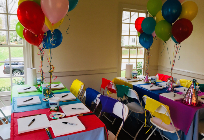 Indoor birthday paint party setup with colorful tables, balloons, and party hats