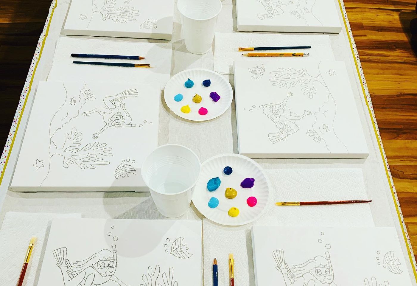 Home dining table set up for a paint party with pre-sketched canvases, paint palettes, brushes, and water cups